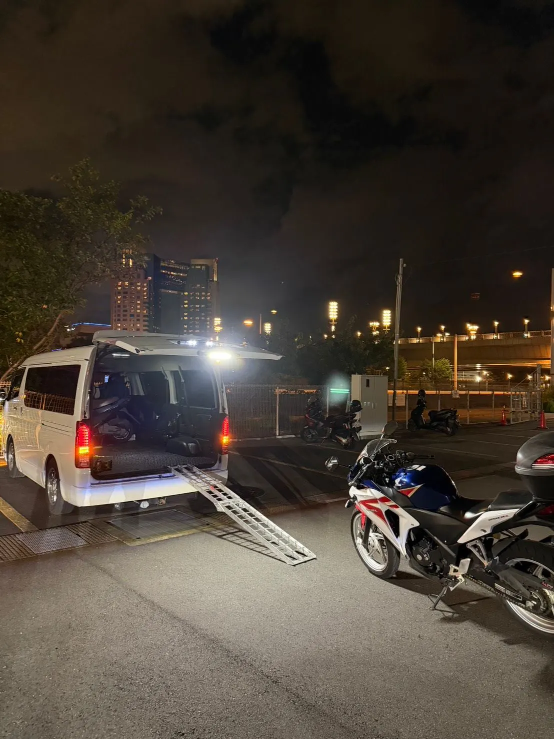 夜の駐車場で、バンの荷室にラダーを掛け、そばにバイクを止めている
